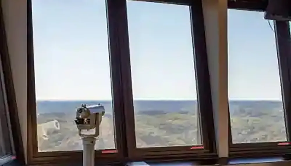 The image shows a coin-operated telescope positioned in front of large windows with a panoramic view of a coastal landscape