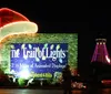 The image shows a festive entrance to The Trail of Lights with a large brightly-lit sign a decorated structure that resembles a Santa hat and a tower adorned with colorful lights in the background at night