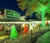 The image shows a festive entrance to The Trail of Lights with a large brightly-lit sign a decorated structure that resembles a Santa hat and a tower adorned with colorful lights in the background at night