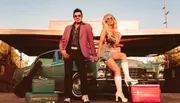 A man and a woman dressed in retro attire pose confidently in front of a vintage car with old-fashioned luggage nearby.