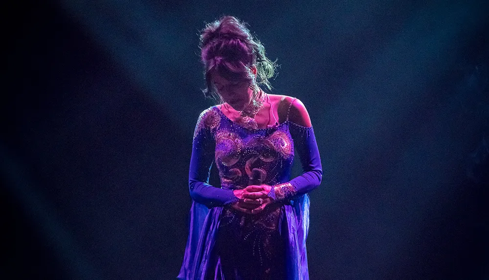 A performer in a blue and purple costume stands with her head bowed under a spotlight on a dark stage