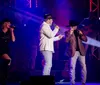 Two men and a woman in cowboy hats are performing on stage with colorful lights and a musician in the background