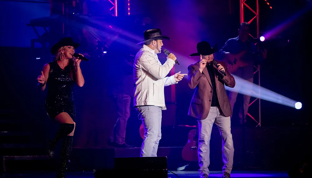 Two men and a woman in cowboy hats are performing on stage with colorful lights and a musician in the background