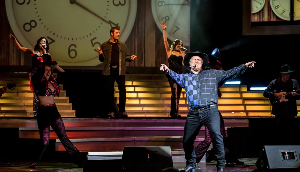 A performer in a cowboy hat is exuberantly singing on stage with musicians and dancers in the background all set against an oversized clock backdrop