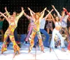 A group of performers in vibrant retro-themed costumes are singing and dancing on stage with a dynamic backdrop that suggests a lively musical atmosphere