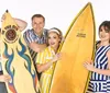 Four people are joyfully posing together with a giant yellow surfboard against a white background