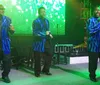 Three men wearing shiny blue jackets are singing into microphones on stage