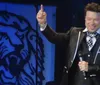 A man in a suit and bow tie is smiling and pointing upwards with one hand while holding a microphone in the other standing in front of a backdrop with a lion design