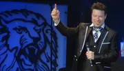 A man in a suit and bow tie is smiling and pointing upwards with one hand while holding a microphone in the other, standing in front of a backdrop with a lion design.