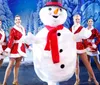 A person in a snowman costume is surrounded by smiling dancers in festive outfits on a winter-themed stage