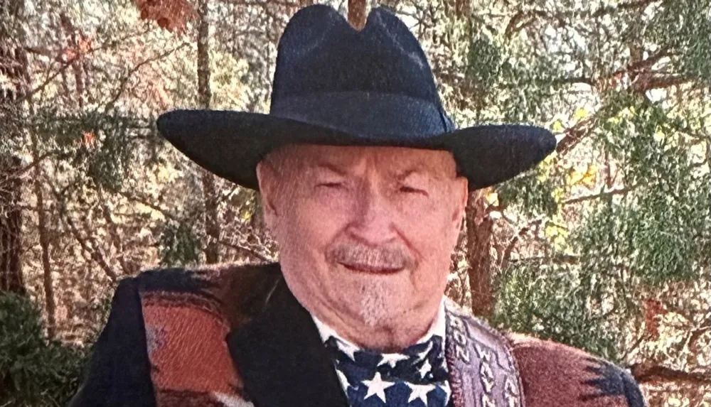 An elderly man with a mustache wearing a cowboy hat and a bandana with an American flag pattern smiles in front of a wooded background