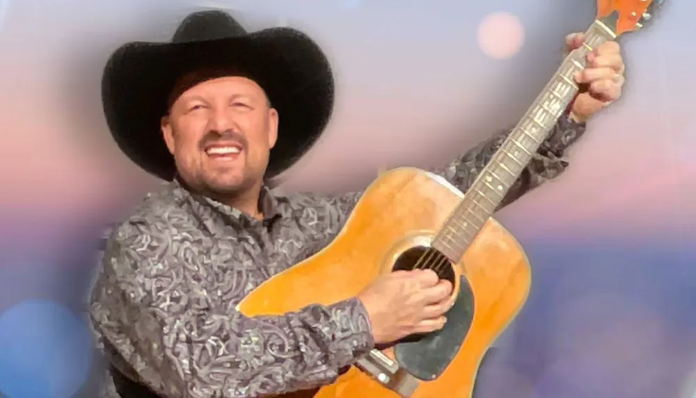 A smiling man wearing a cowboy hat plays an acoustic guitar against a softly blurred background