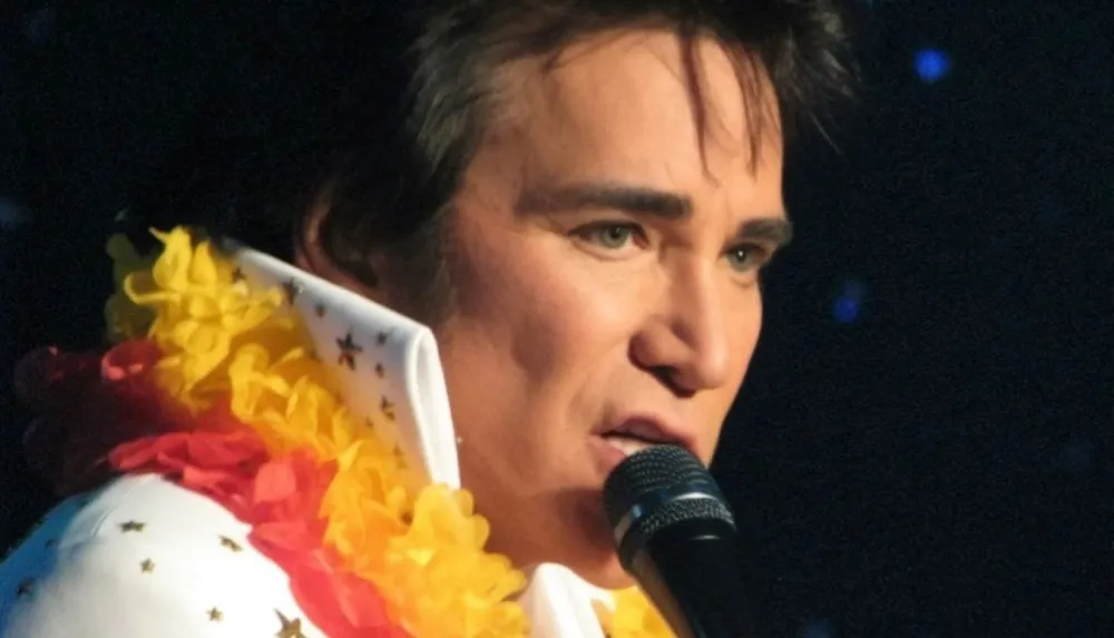 A person is performing onstage with a microphone dressed in an Elvis Presley-inspired outfit adorned with a colorful lei