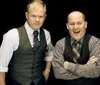 Two men in vests and ties are posing confidently against a black background