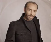 The image shows a middle-aged man with facial hair, wearing a dark coat, and giving a subtle smile against a sepia-toned background.