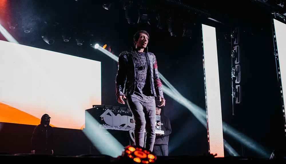 A performer is standing onstage with a confident pose flanked by bright lights and large screens with another person visible in the background at a DJ booth