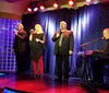 Three singers are performing on stage while a musician accompanies them on a keyboard against a backdrop of a starry curtain and red stage lights