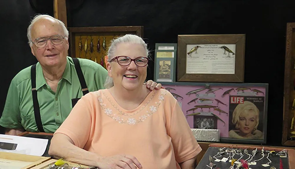 An older couple is smiling for the camera inside a room that displays various items including framed pictures and what appear to be collectibles or crafts on the table in front of them