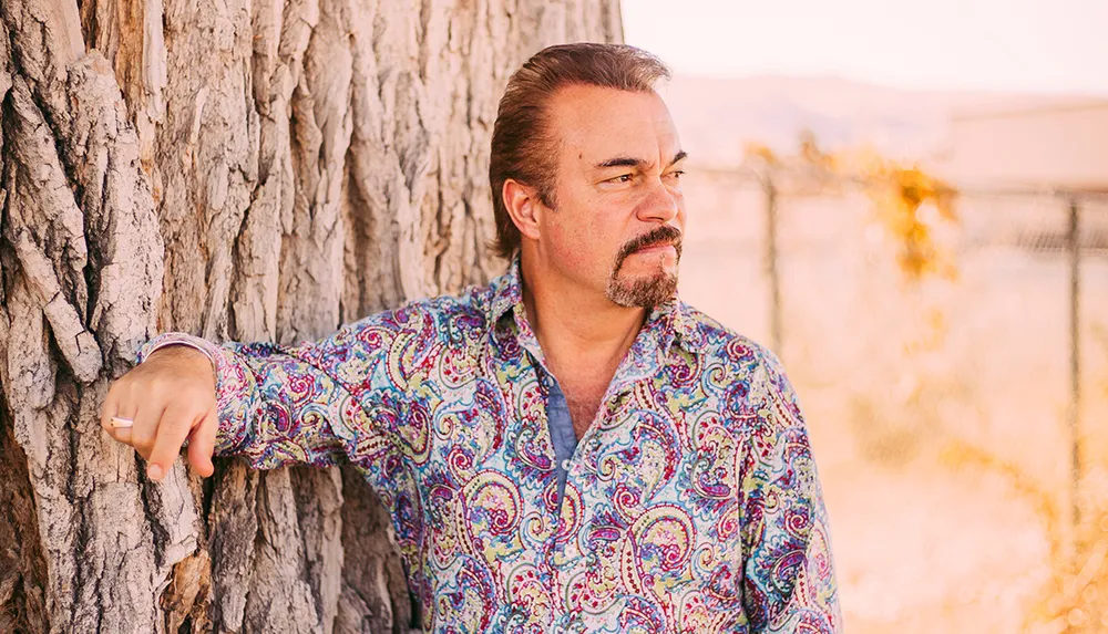 A man with a mustache wearing a colorful paisley shirt leans against a tree gazing pensively into the distance