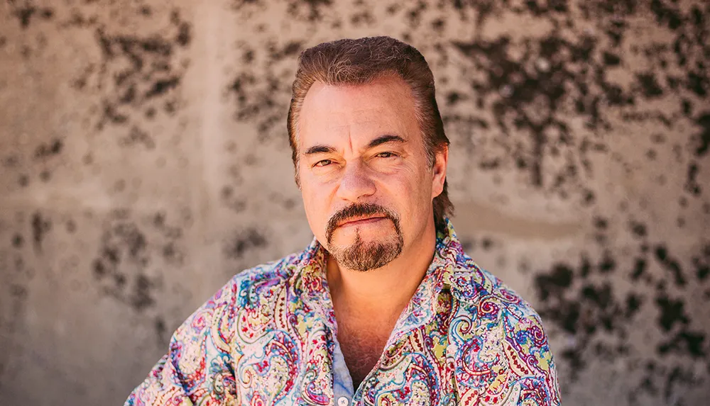 A man with a mustache wearing a colorful shirt offers a half-smile against a textured backdrop