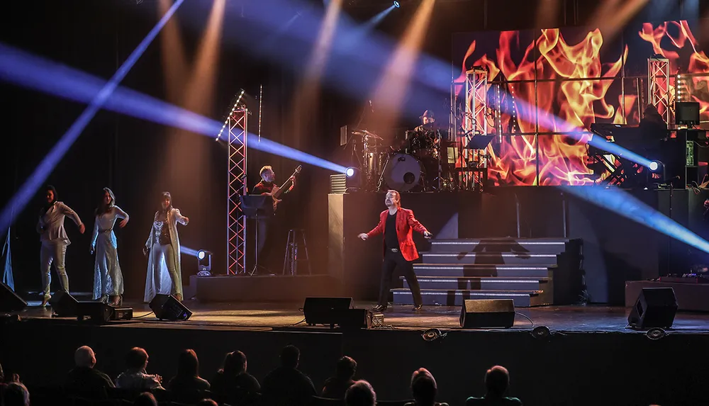 A performer in a red jacket is singing on stage with backup vocalists and musicians under dramatic lighting in front of an audience