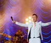 A man in a suit and tie stands on stage with outstretched arms smiling and appearing to engage with an audience under stage lighting