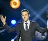 A man in a suit and tie stands on stage with outstretched arms smiling and appearing to engage with an audience under stage lighting