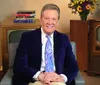 A smiling person is seated with their hands clasped over their knees wearing a blue velvet jacket and paisley tie with books and a vintage television set in the background