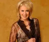 A smiling woman with blonde hair is wearing a brown and gold lace blouse with a warm textured background behind her