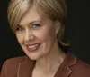 A smiling woman with blonde hair is wearing a brown and gold lace blouse with a warm textured background behind her