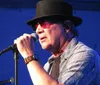 A musician wearing sunglasses and a hat performs into a microphone with musical equipment in the background