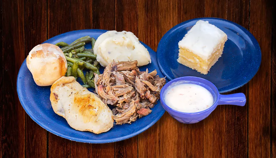 The image shows a hearty meal with grilled chicken breast, pulled pork, green beans, mashed potatoes with gravy, a dinner roll, a square of cake, and a cup of soup, served on a blue plate set against a wooden background.