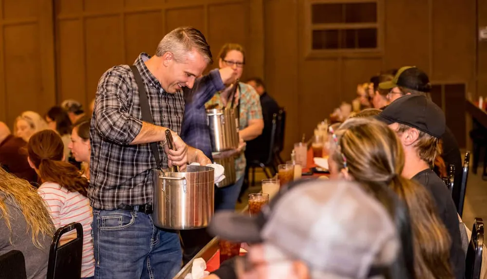 A man is serving food from a large pot to people sitting at banquet tables in a crowded room