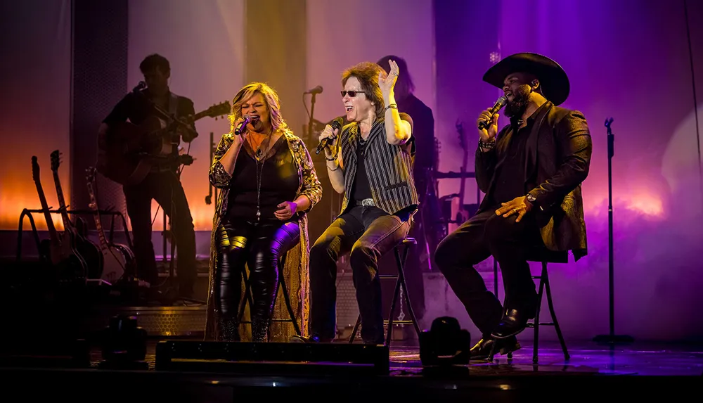 Three vocalists are performing on stage with a guitarist in the background illuminated by warm stage lighting