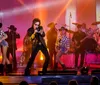 A performer sings passionately on stage flanked by musicians and dancers against a backdrop of warm stage lighting